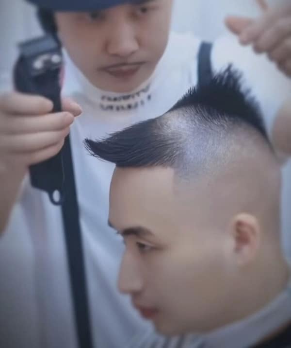 A tragic haircut fail showing a barber cutting a man's hair, leaving a massive bald spot on the top of the head with a tiny, isolated tuft of spiky hair pushed forward at the very front hairline.