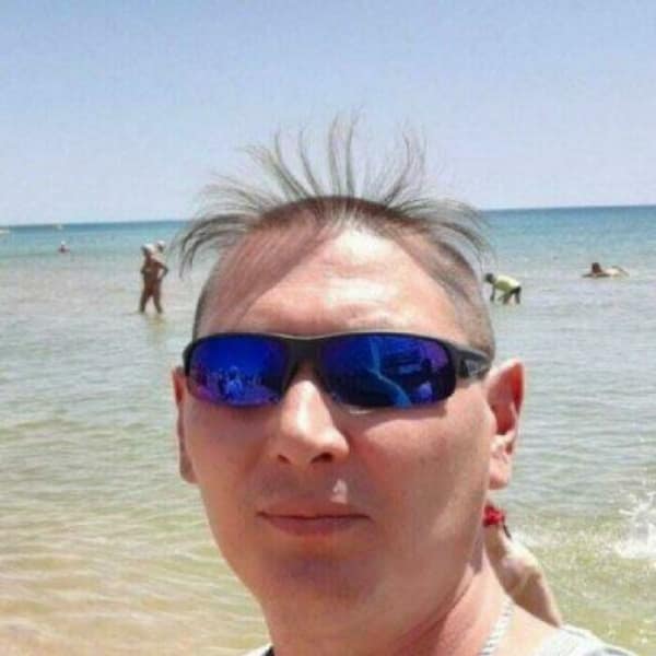 hilarious beach selfie showcasing a legendary bad haircut, where a man has a completely shaved head except for a sparse, wispy fringe that is standing straight up into the air like sparse grass.