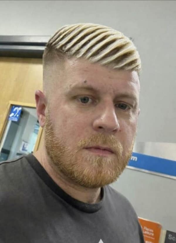 An intensely geometric bad haircut featuring a man with a tight fade on the sides and top hair heavily styled and bleached into deeply grooved, rigid blonde waves that look exactly like dried ramen noodles.