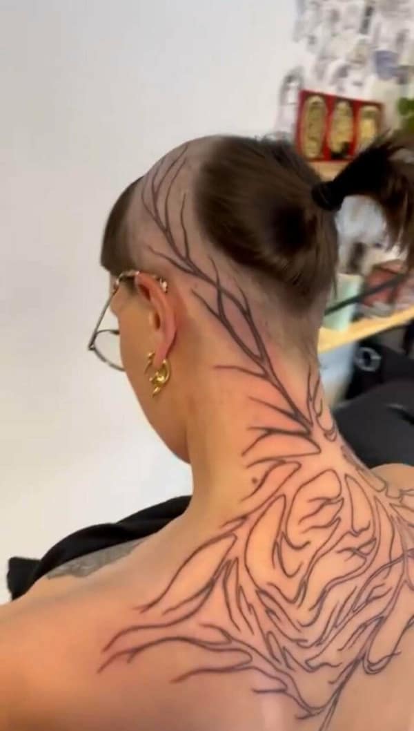 wildly experimental bad haircut showing a person with an entirely shaved back of the head, leaving sharp, geometric lines of hair completely blended into a massive, jagged black line tattoo covering their neck and back.