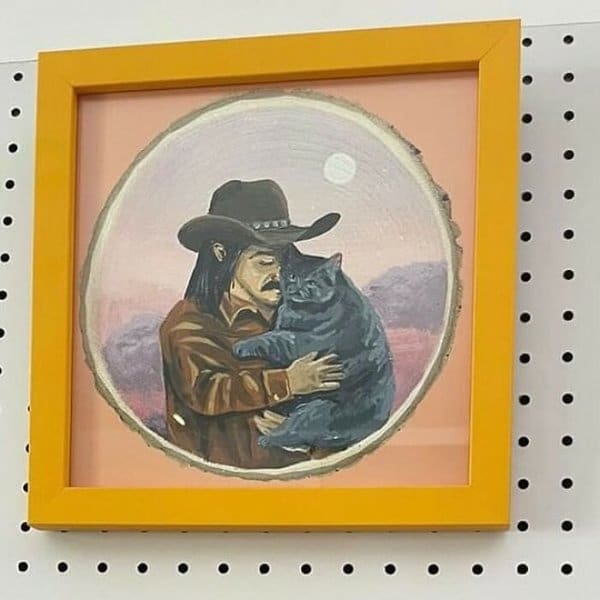 heartwarming and niche piece of amateur art: a circular painting in a bright yellow frame depicting a mustachioed man in a cowboy hat tenderly hugging a large, stoic grey cat.
