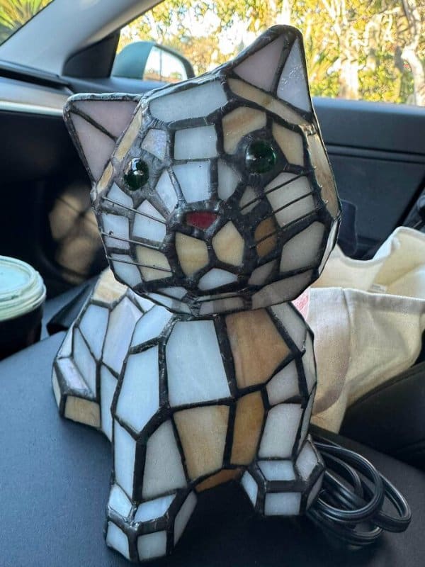 A unique funny thrift store find featuring a desk lamp shaped like a chunky, seated cat made entirely of stained-glass panels in white and tan with glowing green marble eyes.
