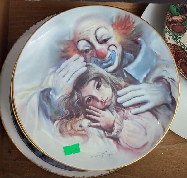 vintage decorative porcelain plate depicting a melancholic clown with blue face paint tenderly cradling a small girl with a single tear on her cheek.