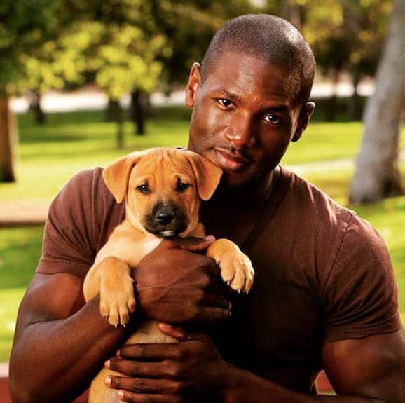 Muscular man tenderly cradling tiny tan puppy outdoors in green park setting