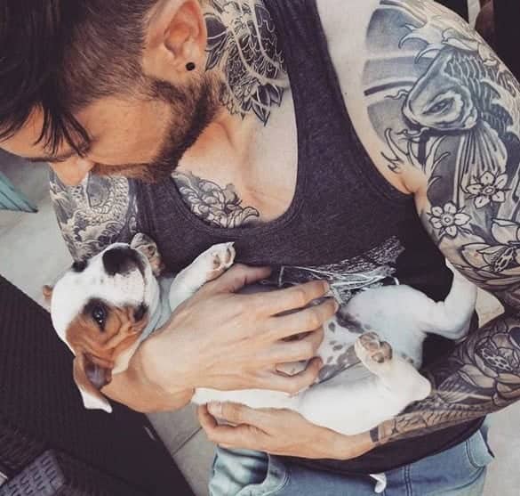 Heavily tattooed muscular bearded man tenderly kissing tiny bulldog puppy close-up