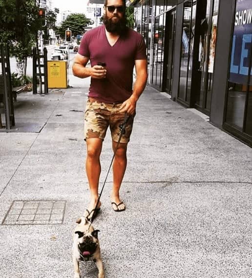 Big bearded man in camo shorts strolling city block with small pug