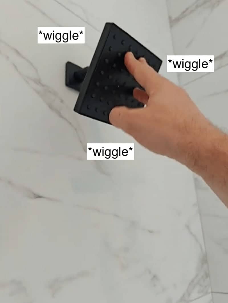 Hand shaking a loose black square shower head against a white marble tiled bathroom wall.