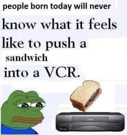 A sad Pepe the frog next to a peanut butter sandwich and a retro video cassette player. A funny classic meme mourning that new generations will never know the bizarre urge to push a sandwich into a VCR.