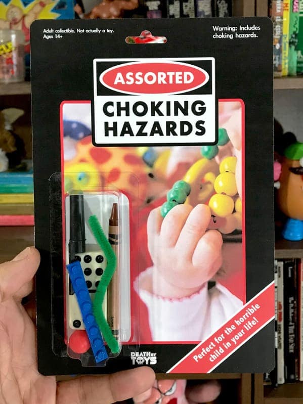 dark humor death by toys set titled "ASSORTED CHOKING HAZARDS," labeled as "Perfect for the horrible child in your life!". The bubble contains a random collection of dangerous small items, including a die, a crayon, a pipe cleaner, and a Lego brick.