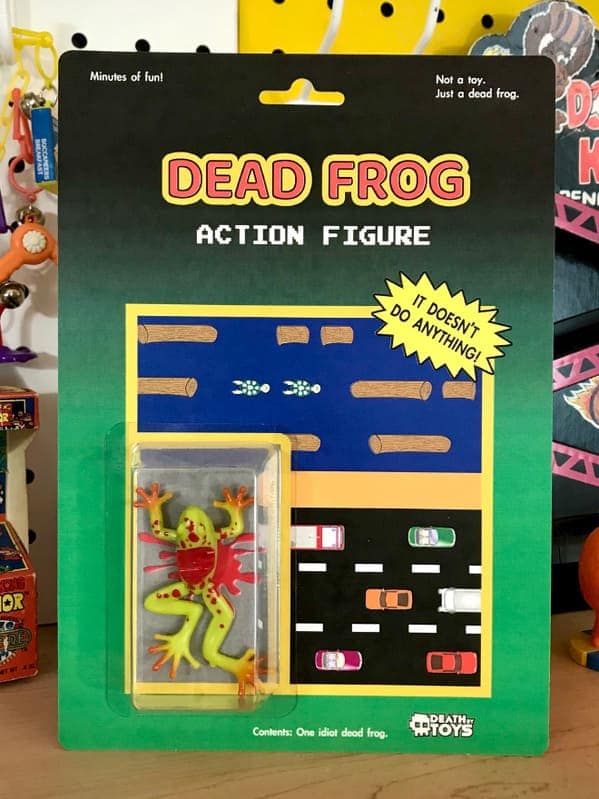 A morbid funny fake action figure titled "DEAD FROG," featuring a flattened, green plastic frog with red blood splatters. The card art parodies the arcade game Frogger, showing the frog crushed on a road full of cars with the tagline: "IT DOESN'T DO ANYTHING!".