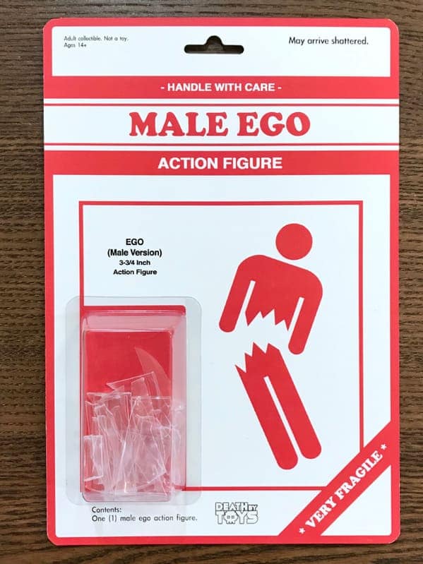 death by toys piece titled "MALE EGO ACTION FIGURE". The packaging contains only shattered shards of clear plastic, labeled "VERY FRAGILE" with a warning that it "May arrive shattered".