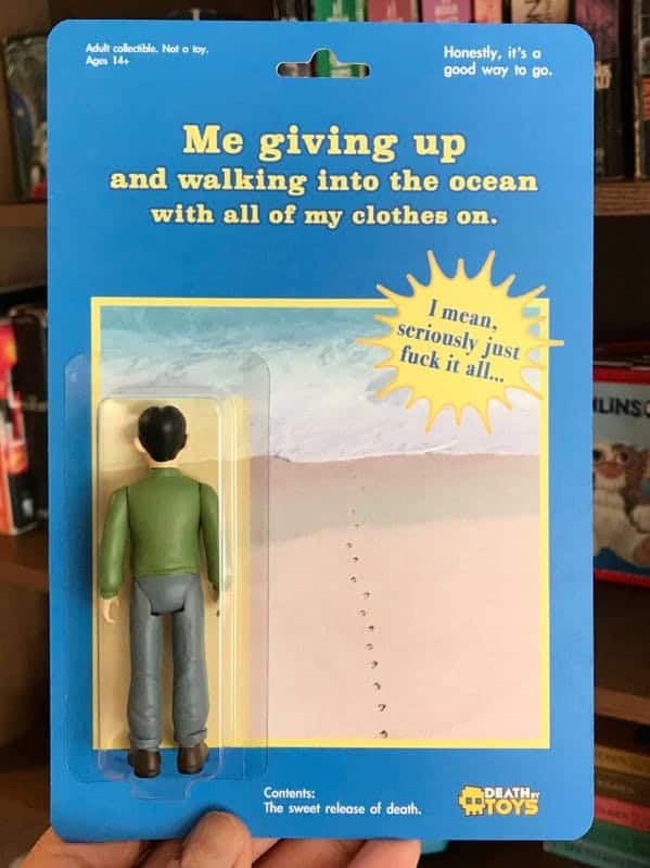 melancholic death by toys piece titled "Me giving up and walking into the ocean with all of my clothes on." The figure is shown from behind, dressed in a green shirt and grey pants, facing a card back of a desolate beach with the dark caption: "I mean, seriously just [censored] it all..."
