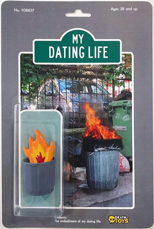 A literal interpretation of a "dumpster fire" as a funny fake action figure titled "MY DATING LIFE." The toy consists of a small grey trash can with a bright orange and yellow plastic flame protruding from the top, packaged over a photo of an actual burning dumpster.
