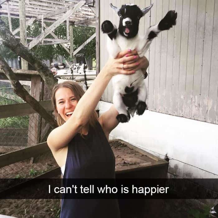 A woman holding up a tiny baby goat that looks incredibly happy with its mouth open.