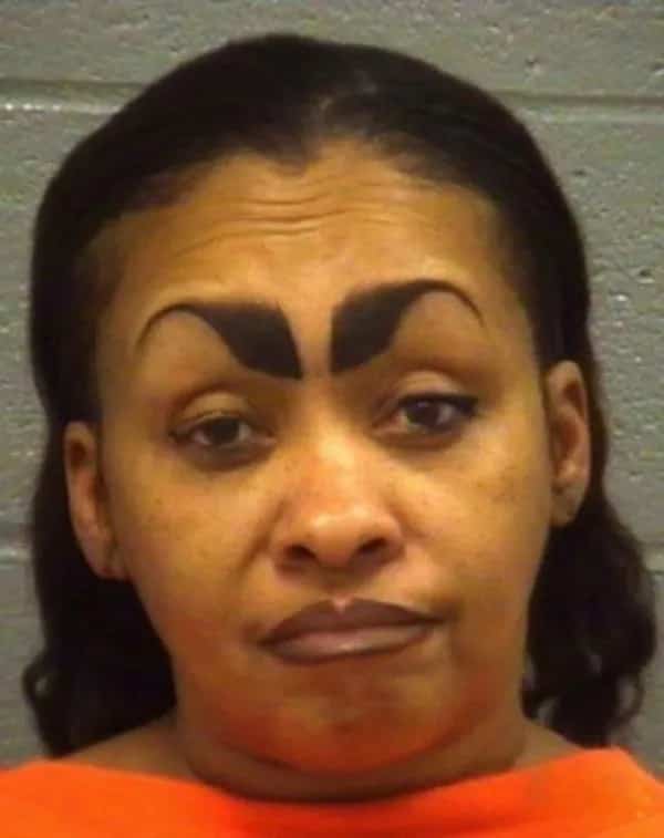 Mugshot of a woman featuring oversized thick black eyebrows with a sharp triangular inner point.