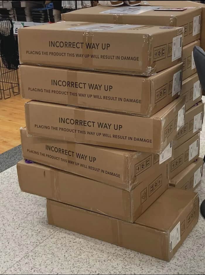 A quintessential funny fail of warehouse logistics: a stack of large cardboard boxes where every single box is printed with the warning "INCORRECT WAY UP" in bold letters, and the entire stack is oriented exactly in that forbidden position.