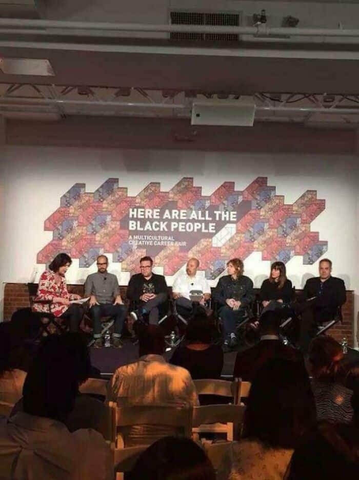 An ironic and poorly staged funny fail at a "Multicultural Creative Career Fair." The panel sits beneath a massive wall graphic that reads "HERE ARE ALL THE BLACK PEOPLE," while every single member of the seven-person panel appears to be white.