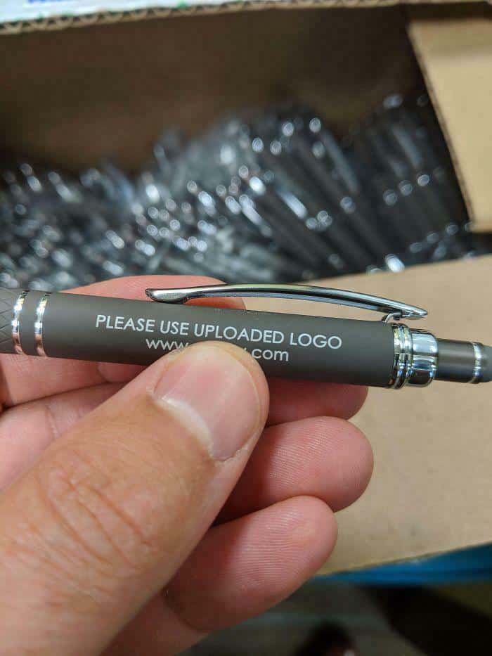 A customized merchandise funny fail where a silver and grey promotional pen is engraved with the literal instruction "PLEASE USE UPLOADED LOGO" and a website URL instead of the actual company logo.