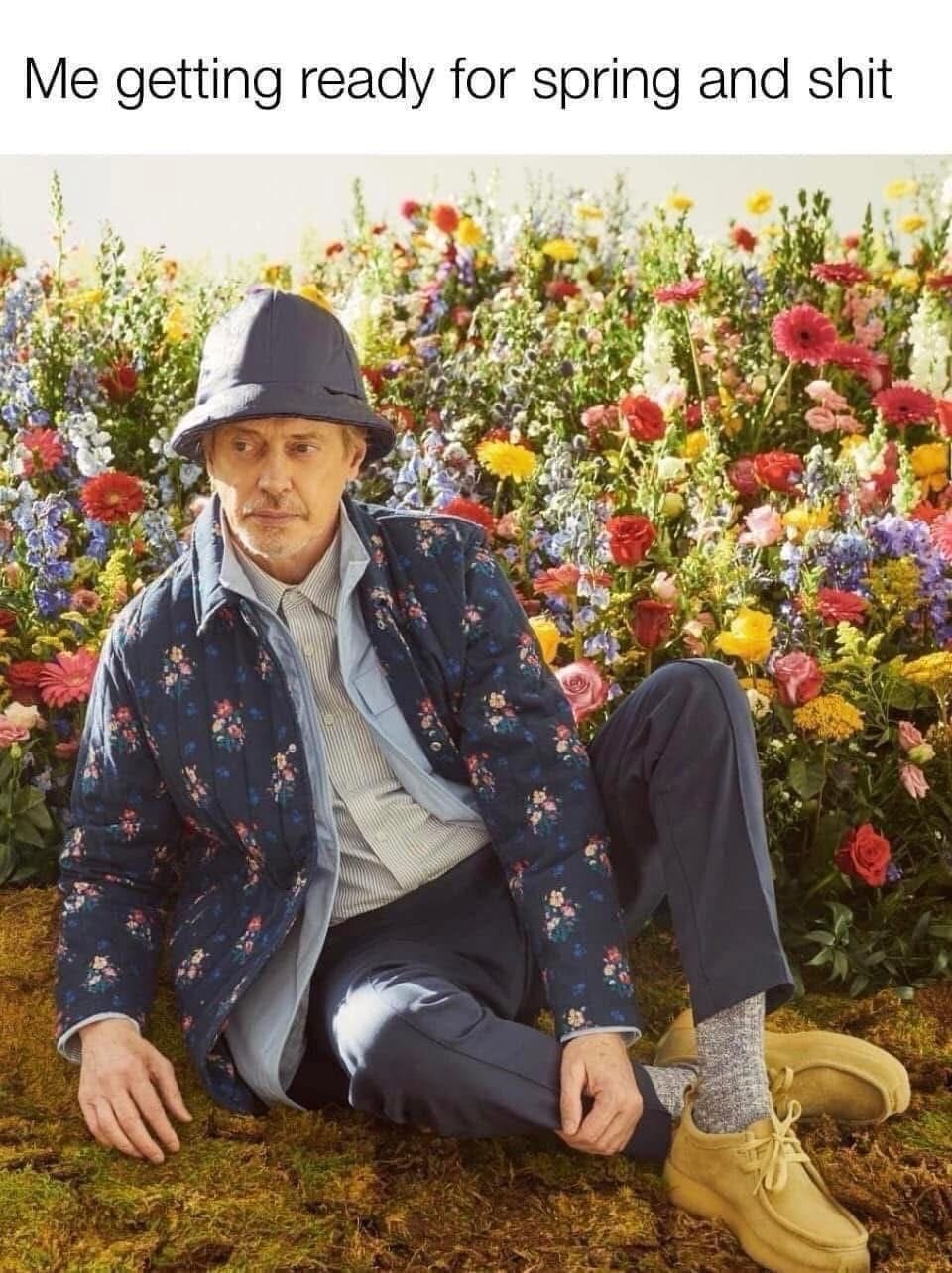 Actor Steve Buscemi wearing a floral jacket and bucket hat sitting in a field of flowers.