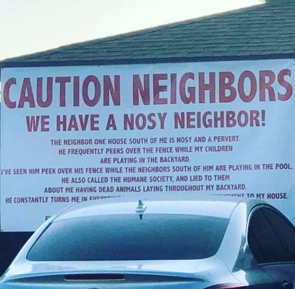 The ultimate public shaming: a massive white banner with red text hanging from a building that screams, "CAUTION NEIGHBORS WE HAVE A NOSY NEIGHBOR!" The sign goes on to accuse a specific neighbor of being a "pervert" who peeks over fences and calls the Humane Society with lies about dead animals.