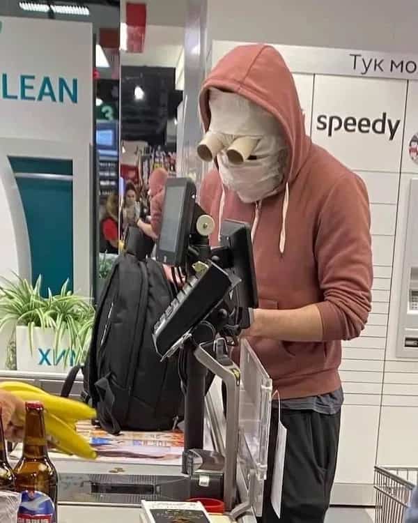 Person at a grocery checkout wearing a bizarre mask made of bandages and cardboard tube eyes.