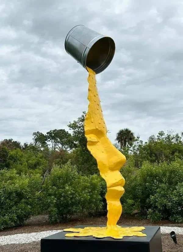 Large outdoor sculpture featuring a giant silver bucket pouring yellow paint that forms a human face.