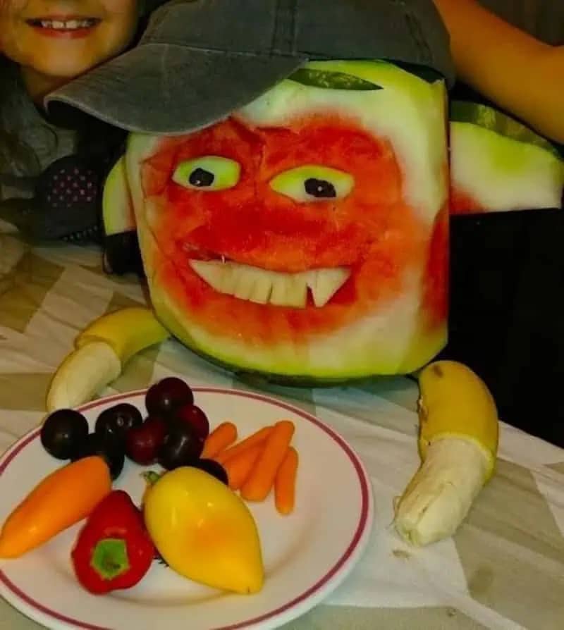 Creative fruit art of a watermelon face with banana arms and a grey baseball cap.