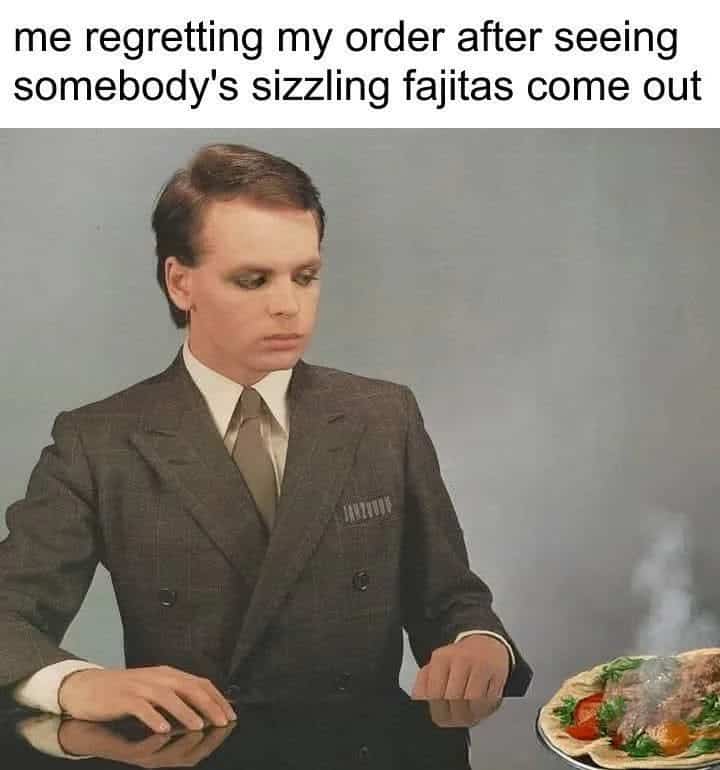 A man in a suit with dark eye makeup looking regretfully at a plate of sizzling fajitas.