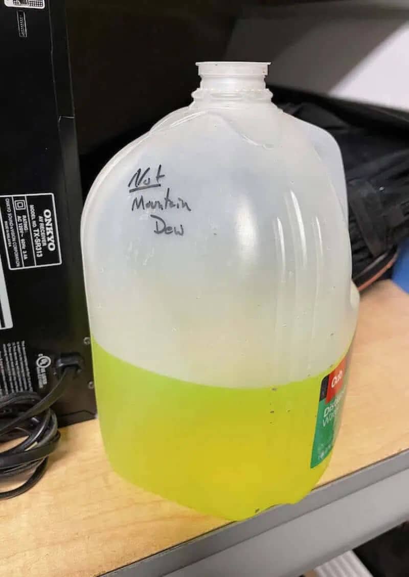 A plastic milk jug filled with neon green liquid labeled not mountain dew sitting on a wooden shelf.