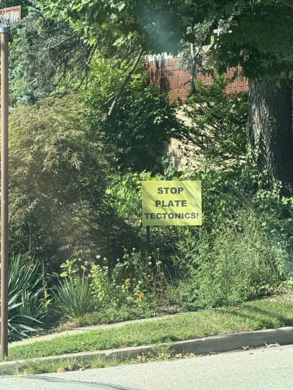An absurdly ambitious yellow lawn sign stands among garden bushes, demanding to "STOP PLATE TECTONICS!" as if geological forces were a local political issue
