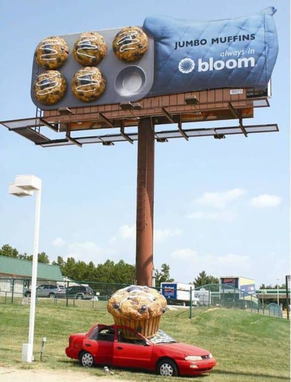 A massive 3D funny sign billboard for "Jumbo Muffins." As part of a clever guerrilla marketing campaign, a giant, realistic muffin has seemingly "fallen" from the sign and crushed the roof of a real red car parked on the lawn below.
