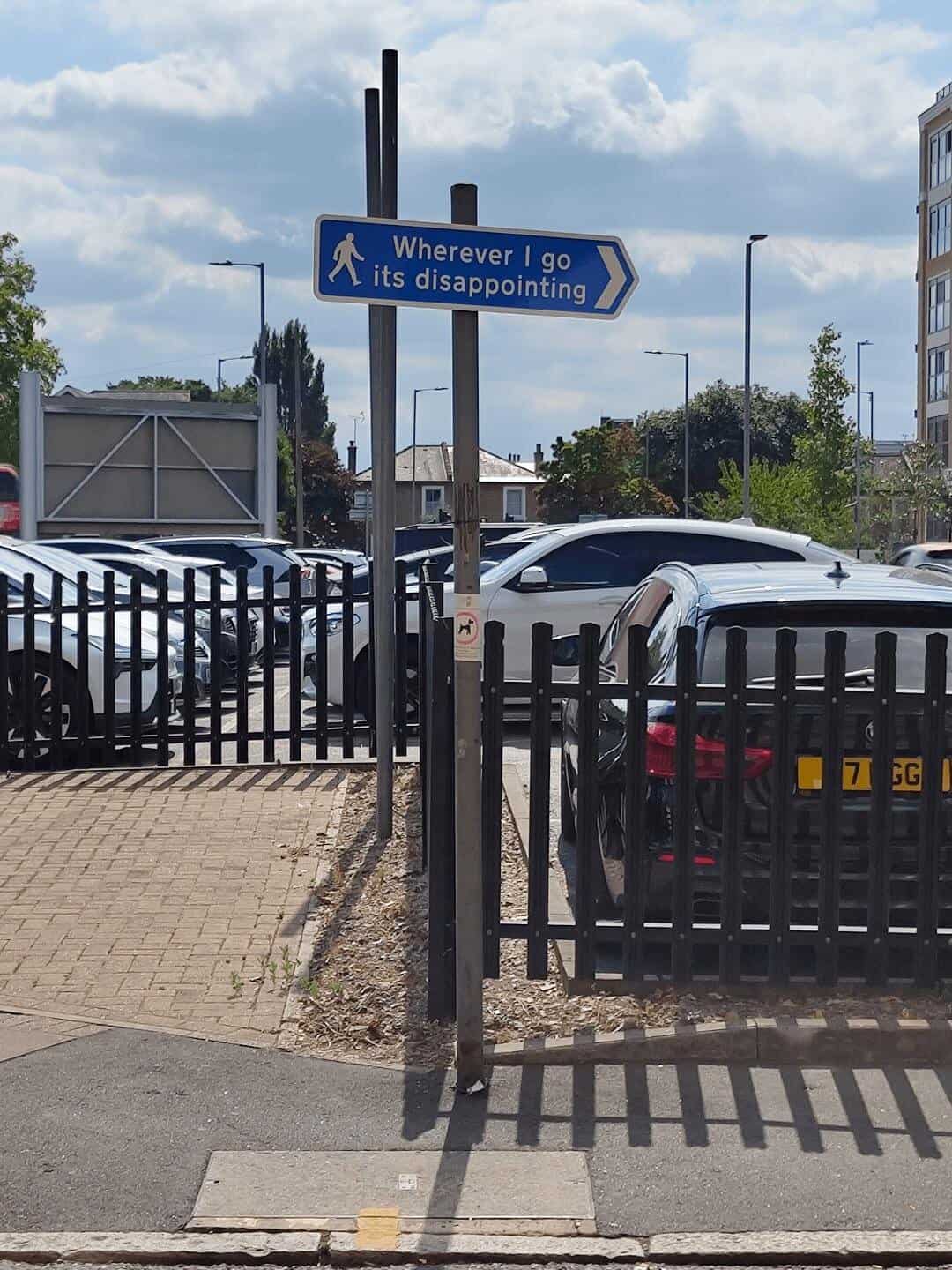A blue pedestrian sign in a parking lot offers a dose of deep cynicism, directing travelers toward a path labeled "Wherever I go its disappointing."