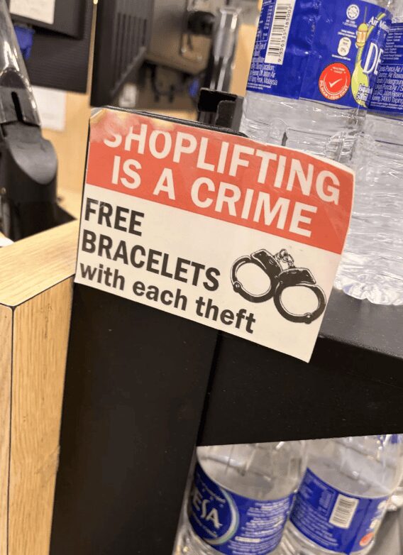 A snarky funny sign at a retail checkout counter. The header "SHOPLIFTING IS A CRIME" is followed by a sarcastic offer of "FREE BRACELETS with each theft," featuring an illustration of a pair of metal handcuffs.