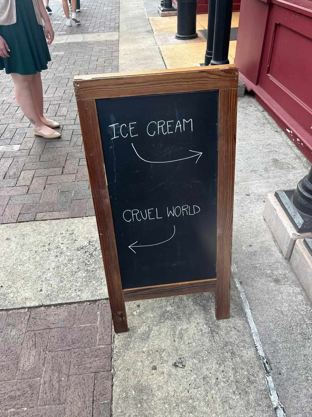 chalkboard A-frame on a sidewalk offers a simple choice to passersby, with an arrow pointing right toward "ICE CREAM" and an arrow pointing left back toward the "CRUEL WORLD."