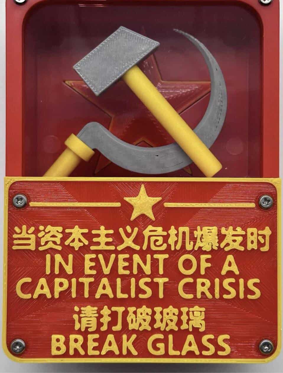 A satirical emergency box features a hammer and sickle behind glass with a bilingual red plaque stating, "IN EVENT OF A CAPITALIST CRISIS BREAK GLASS."