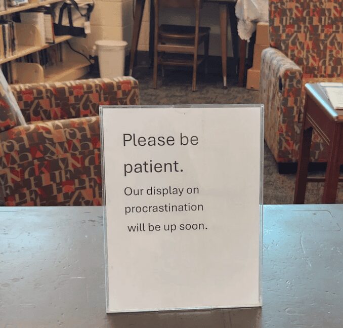 A self-referential funny sign sitting on a counter in a clear plastic stand. It asks for the viewer’s indulgence with the message: "Please be patient. Our display on procrastination will be up soon."