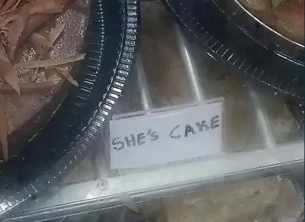 grocery store find where a small sign in a refrigerated case reads "SHE'S CAKE," a funny spelling mistake that likely meant to label a "cheesecake" or "sheet cake" but ended up personifying the dessert.