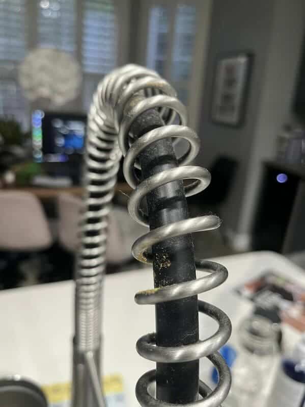 A close-up of a kitchen faucet's metal spring coil covered in dust and food particles.