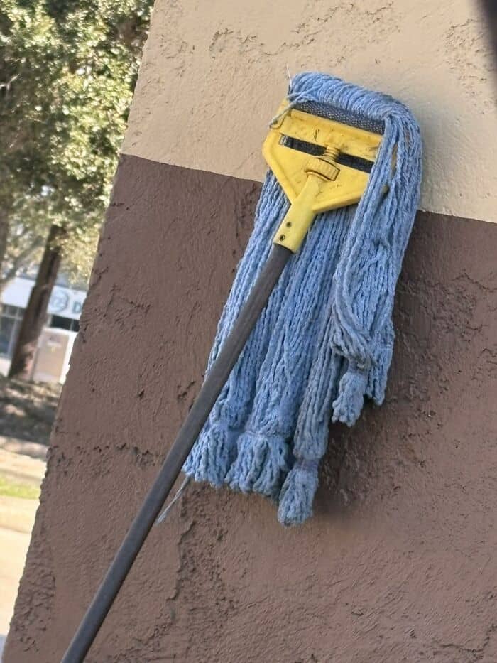 inanimate objects with faces showing a blue string mop leaning against a wall, where the yellow plastic clamp creates a tired, exhausted face with long blue dreadlocks.
