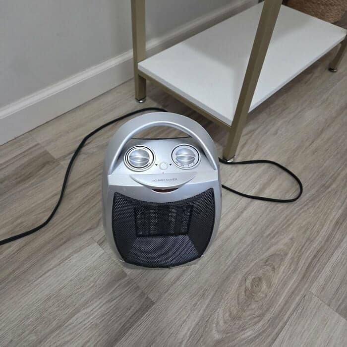 delightful find of inanimate objects with faces showing a small silver space heater on a floor, using its two round control dials and black grill to form a wide, friendly, frog-like smile.