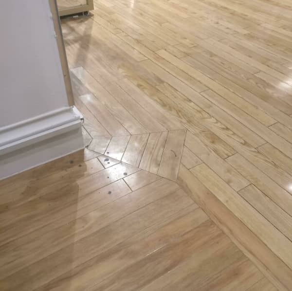 Light wood flooring with planks meeting at nonsensical, jagged angles near a white wall baseboard.