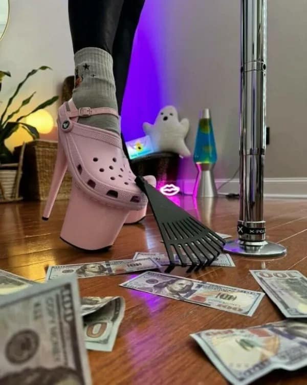 A foot wearing a massive pink Croc designed as a platform high heel shoe stepping on a tiny rake surrounded by cash. Bizarre cursed footwear in this meme dump.