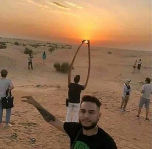 A surreal desert scene where a man appears to have elongated, elastic arms that reach high into the sky to perfectly "pinch" and frame the setting sun.