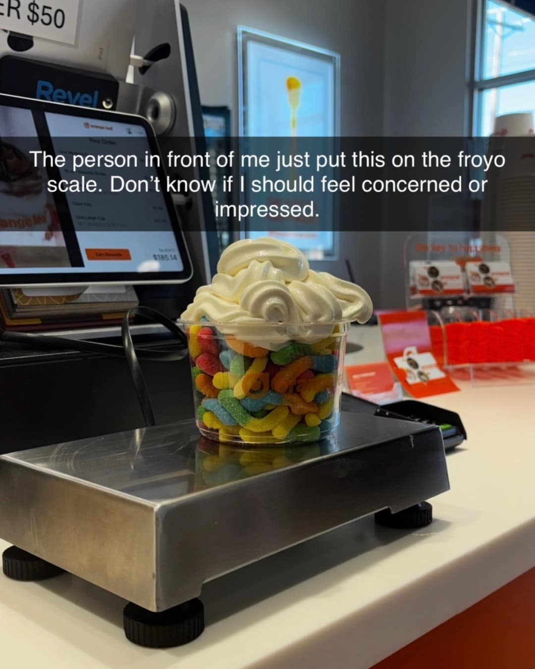 An impressive yet concerning meme dump photo of a frozen yogurt cup packed to the brim with colorful gummy worms and topped with a heavy swirl of vanilla froyo sitting on a checkout scale.
