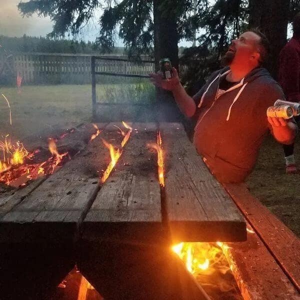chaotic meme dump image of a man standing at a party next to a wooden picnic table that has been set on fire. He is holding several beer cans and looking up in a moment of frantic, drunken energy.
