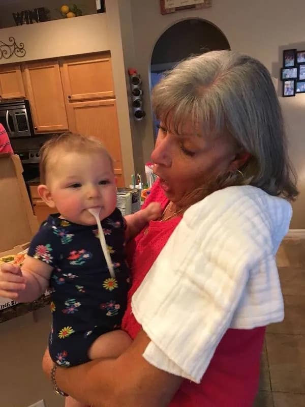 perfectly timed meme dump photo of a mother holding a baby. The baby is spitting up a stream of milk so solid and perfectly shaped that it creates an optical illusion of a white plastic spoon being held in its mouth, much to the mother’s visible shock.