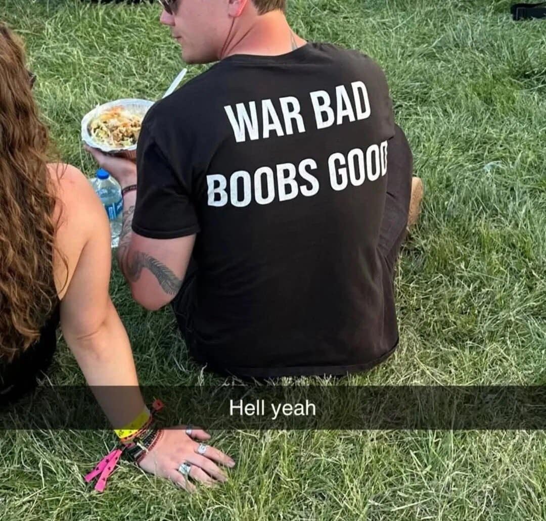 A guy sitting on the grass wearing a black t-shirt with the profound and simple political statement War Bad Boobs Good. A hilarious meme to include in a chaotic meme dump.