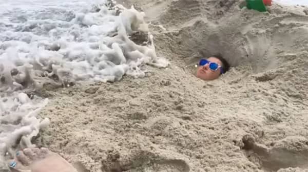 A person buried in beach sand as a large ocean wave begins to crash over.