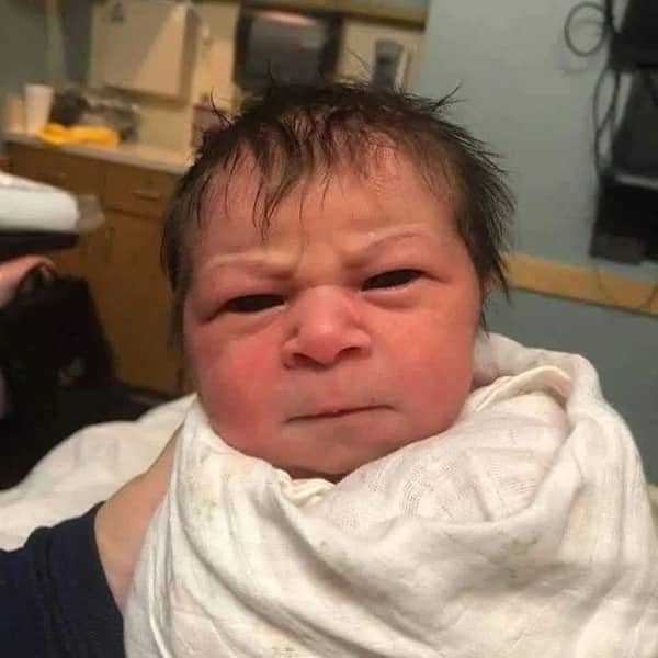 Newborn baby with messy hair and an annoyed scowl looking like they missed their retirement.