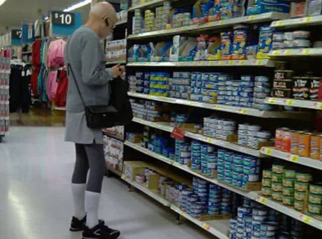 Bald man in the canned goods aisle wearing a grey skirt, leggings, and white socks.